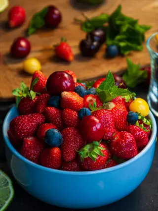 a blue bowl filled with fresh berries including strawberries, blueberries, cherries, and mint leaves, surrounded by natural sweeteners like fruits on a wooden surface