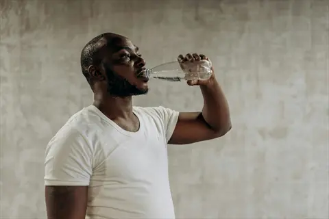a person drinking water from a clear plastic bottle while wearing a white t-shirt against a plain gray background