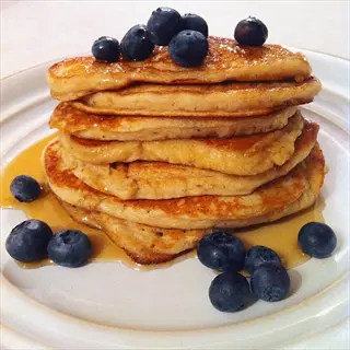 a tall stack of almond flour pancakes topped with fresh blueberries and drizzled with maple syrup on a white plate