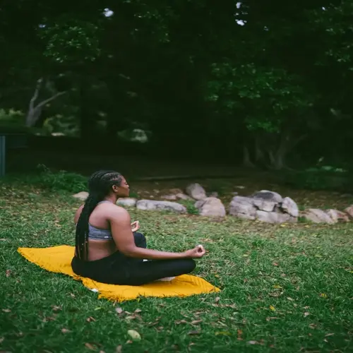 a woman meditating in a calm park, sitting in lotus position on a yellow cloth surrounded by trees and rocks