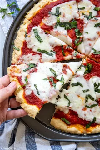 almond flour pizza crust topped with tomato sauce, melted mozzarella, and fresh basil, with a slice being lifted from the pan (watermark: cinnamon)