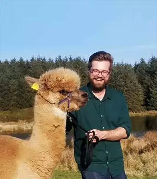 alpaca therapy session: man with beard and glasses smiling while holding the lead of a friendly light brown alpaca in sunny outdoor setting with lake and evergreen forest