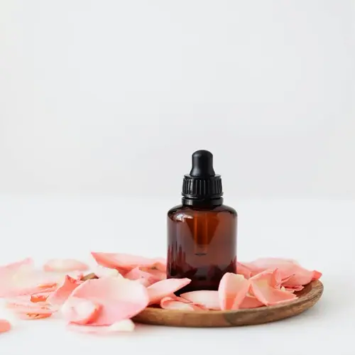 amber glass rose oil bottle with dropper, surrounded by pink rose petals on a wooden tray