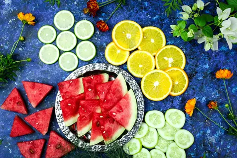 artful arrangement of watermelon cucumber slices with lime, orange, and cucumber on blue background, featuring a silver platter and decorative flowers