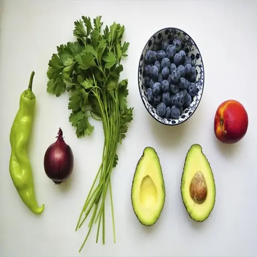 assortment of antioxidant rich foods: green chili pepper, fresh parsley, bowl of blueberries, nectarine, and avocado halves on white surface
