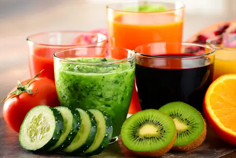 assortment of fresh vegetable juices with accompanying produce: green cucumber blend, purple beet, orange carrot, and red tomato drinks surrounded by sliced veggies and fruits