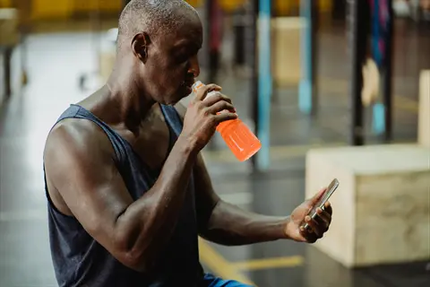 athlete drinking juice from a sports bottle while checking smartphone in a gym
