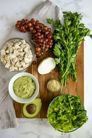 avocado cream dip in a white bowl on a wooden cutting board with fresh ingredients: red grapes, chicken, parsley, avocado, mustard, and lettuce