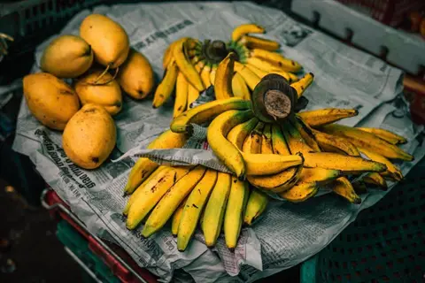 banana bunches and mangoes on newspaper-covered table with visible text; includes banana bunch wooden table elements (table appears non-wooden)