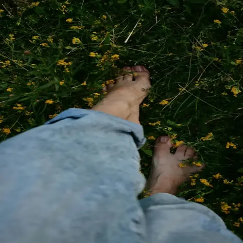 bare feet walking on grassy field with yellow flowers, wearing light blue jeans - first-person perspective