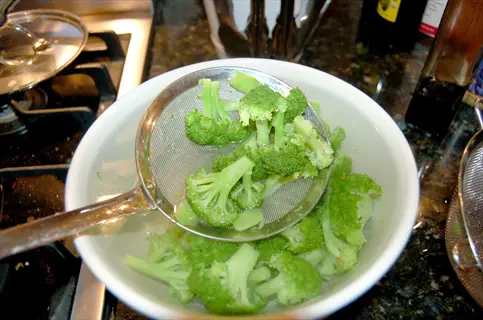 blanching vegetables pot: fresh broccoli florets in a white bowl with a strainer, surrounded by kitchen stove and condiments