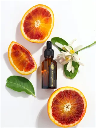 blood orange essential oil bottle surrounded by fresh blood orange slices, whole fruit, citrus blossoms and green leaves on white background