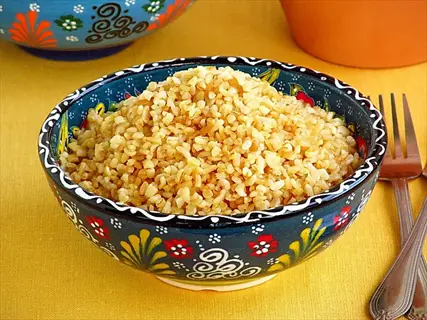bowl of cooked bulgur wheat in decorative ceramic dish with floral patterns, forks and background bowls on yellow tablecloth