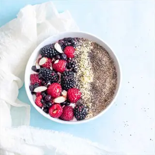 bowl of greek yogurt topped with mixed berries, almonds, and hemp seeds on light blue background