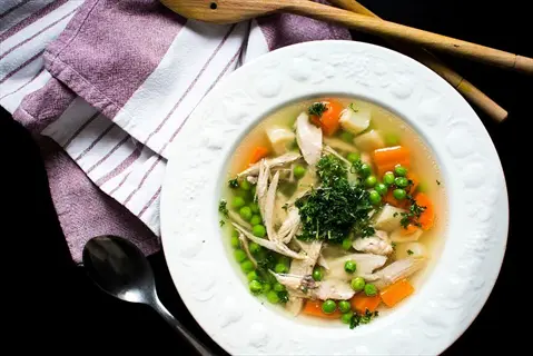 bowl of homemade bone broth soup with shredded chicken, carrots, peas, and parsley, served with wooden spoons and a striped cloth napkin