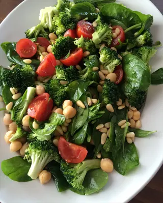 broccoli spinach salad with cherry tomatoes, chickpeas, and pine nuts. fresh vegetable medley featuring leafy greens and cruciferous florets in a white bowl