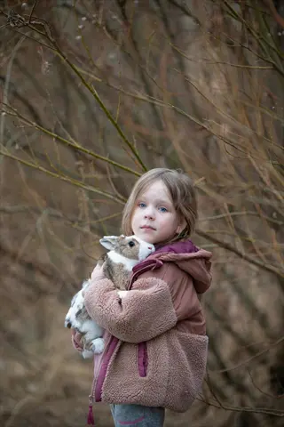 child holding rabbit: young girl in brown fleece jacket cradling small rabbit outdoors among bare branches