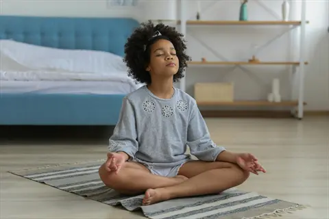 child meditation: young girl in grey sweatshirt meditating in lotus pose on a woven rug in a bright bedroom with blue furniture