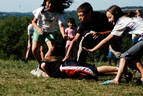 children playing tag on sunny grassy field with one child falling/tagged while others reach to assist or tag
