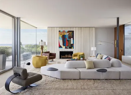 clean living room with minimalist neutral furniture (white sectional sofa, yellow armchairs), abstract art above fireplace, floor-to-ceiling windows overlooking greenery, and sculptural lighting fixtures
