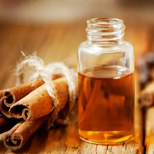 clear glass bottle of golden cinnamon oil beside bundle of cinnamon sticks tied with twine on weathered wooden surface; no text visible