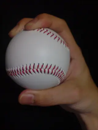 close-up of a baseball gripped in a four-seam fastball pitch grip, with fingers positioned across red stitching seams and thumb underneath, against a dark background
