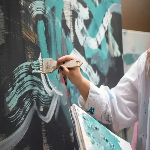 close-up of a person painting art: hand holding paintbrush applying teal and white abstract strokes on dark canvas, paint splatters on white shirt