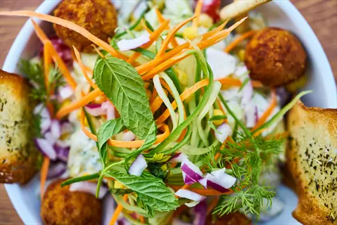 close-up of a vibrant salad bowl featuring crunchy vegetables: shredded carrots, cucumber ribbons, red onion, fresh mint and dill, crispy bread slices, and croquettes