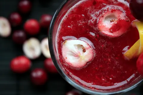 close-up of cranberry juice vitamin a beverage in glass with halved cranberries and lemon slice, surrounded by whole cranberries on dark background
