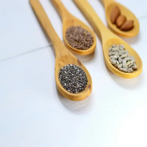 closeup of wooden spoons displaying chia seeds, flax seeds, sunflower seeds, and almonds on white surface
