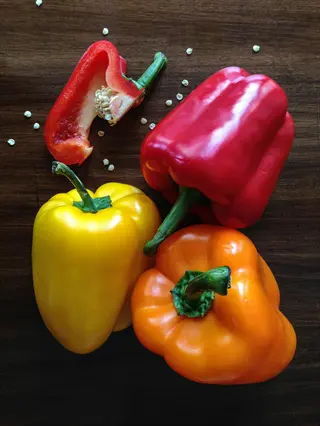 colorful bell peppers: whole red, yellow, and orange peppers with a sliced red pepper and scattered seeds on dark wood surface
