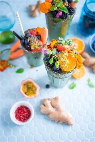 colorful food arrangement: green smoothie glasses adorned with strawberries, blueberries, blackberries, passion fruit, orange slices, carrot sticks, and edible flowers. surrounded by fresh ginger, passion fruit halves, and red spices on blue tiled surface