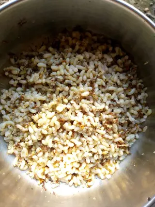 cooked barley grains: a metal bowl filled with light tan elongated grains (barley) mixed with small brown specks, possibly spices or seasonings