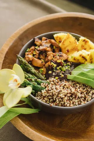 cooked quinoa bowl featuring grilled chicken, asparagus, pineapple, avocado, and sauce. served in a wooden dish with orchid and herb garnish. nutrient-rich grains base