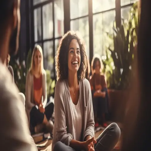 dance therapy group session: diverse individuals sitting in a circle during a discussion in a sunlit, plant-filled room, featuring a smiling woman in a gray cardigan