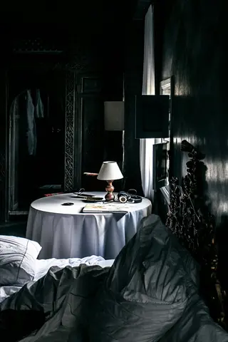 dimly lit dark cool bedroom with ornate carved walls, unmade bed, and bedside table holding a lamp, headphones, and book