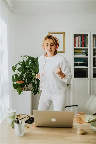 energized woman in white sweatsuit enjoys office music relaxation with pink headphones, dancing by laptop in bright home office with plants and bookshelves