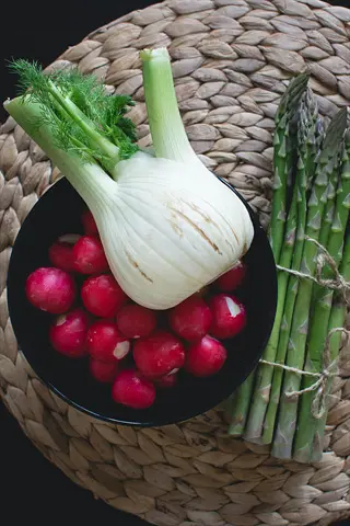 fresh fennel bulb with fronds resting on bowl of radishes, alongside bundled asparagus spears (prebiotic fiber sources similar to garlic/onions) on woven placemat