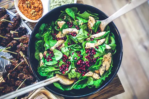 fresh spinach salad in black bowl with chicken, pomegranate seeds, and serving utensils