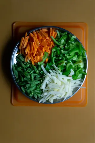 fresh vegetable cutting: carrot, green bell pepper, green beans and potato strips in a metal bowl on an orange cutting board, demonstrating preparation timing