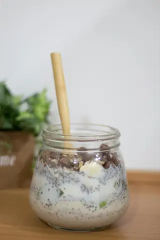 glass jar of layered overnight oats with chia seeds (visible seeds), layered with yogurt/fruit, wooden spoon, and potted plant background