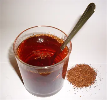 glass of brewed rooibos tea with spoon and dried rooibos leaves pile on white background