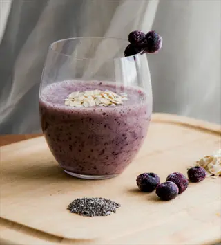 glass of chia smoothie topped with oats and blueberries, with chia seeds and fresh blueberries arranged on a wooden board