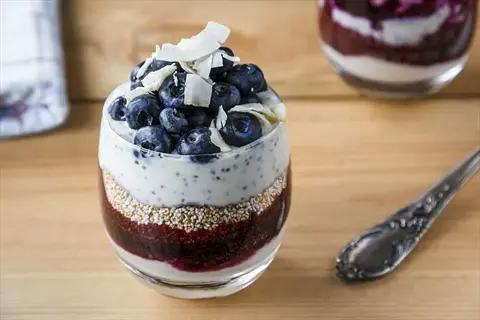 greek yogurt berries parfait: layered with blueberry compote, granola, and coconut flakes in a glass. vegetarian dessert with fresh blueberries and a spoon on a wooden table