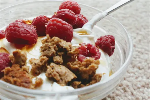 greek yogurt bowl with fresh raspberries, crumbled granola, and honey drizzle in clear glass bowl with spoon on textured beige surface