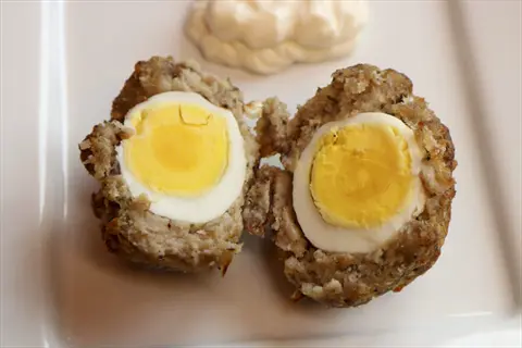 halved scotch eggs showing boiled eggs portion, coated in seasoned meat and breading, served with creamy sauce on white plate