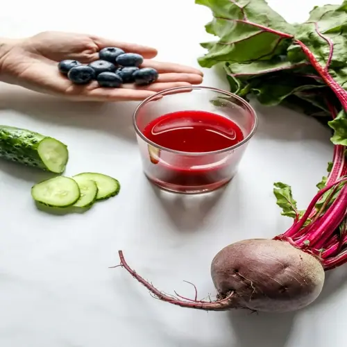 hand holding blueberries, glass of fresh beetroot juice, sliced cucumbers, and whole beetroot with greens on white background
