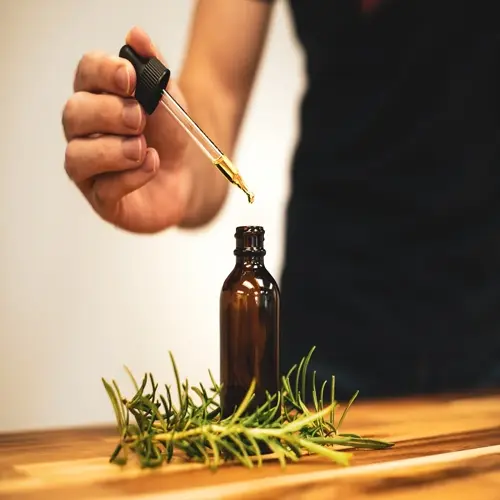 hand holding dropper releasing rosemary essential oil into amber glass bottle with fresh rosemary sprigs on wooden table