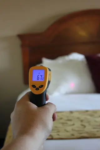 hand holding infrared bedroom thermometer measuring bed temperature (67.0°f display) in cozy bedroom with wooden headboard and white bedding
