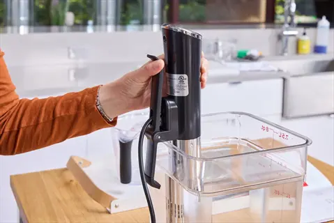 hand placing sous vide machine immersion circulator into water bath container with volume markings (7.5l, top 8) in modern kitchen